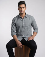 A man wearing a light gray long-sleeve button-down shirt with a chest pocket, posed against a neutral light gray studio background.