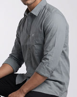 A man wearing a gray long-sleeve button-down shirt with rolled-up sleeves and black pants, shown from the chest down against a light gray background.