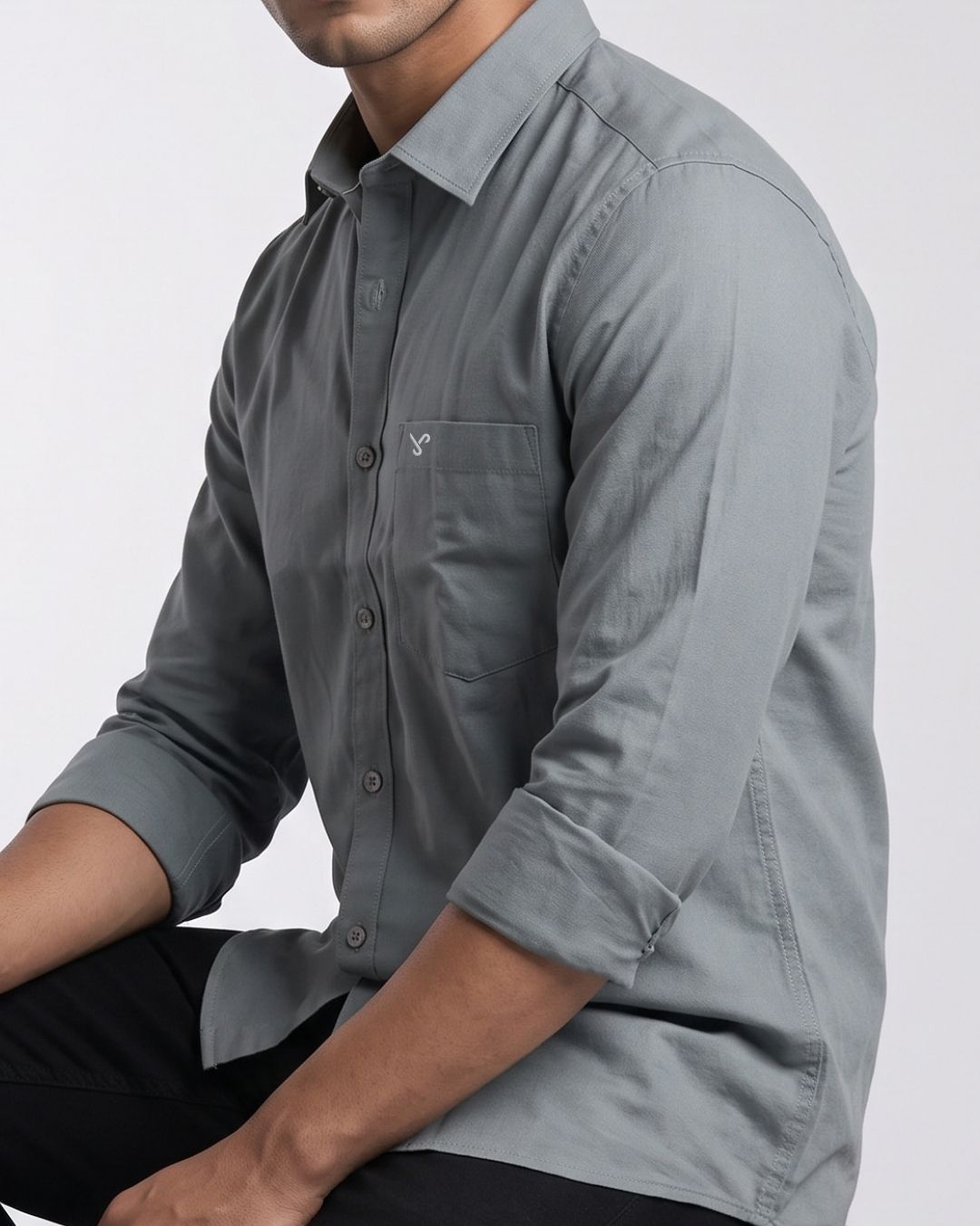 A man wearing a gray long-sleeve button-down shirt with rolled-up sleeves and black pants, shown from the chest down against a light gray background.