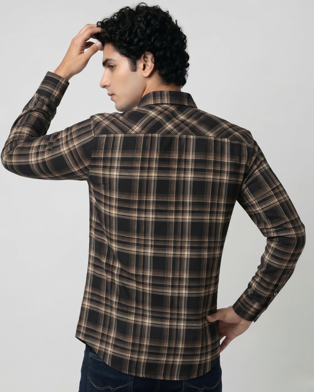 A man wearing a long-sleeve plaid button-up shirt in brown, black, and beige tones, shown from a three-quarters back view against a light gray studio background.