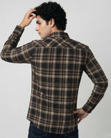 A man wearing a long-sleeve plaid button-up shirt in brown, black, and beige tones, shown from a three-quarters back view against a light gray studio background.