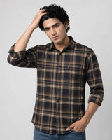 A man wearing a long-sleeve button-up plaid shirt in dark brown, black, and beige, paired with blue jeans, posing against a light gray studio background.