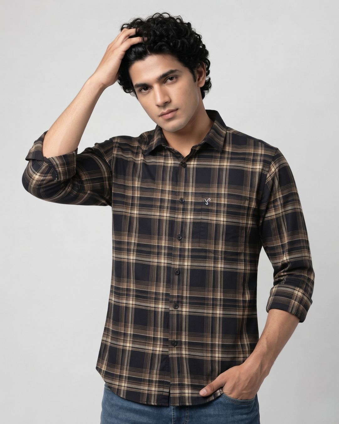 A man wearing a long-sleeve button-up plaid shirt in dark brown, black, and beige, paired with blue jeans, posing against a light gray studio background.