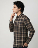A man wearing a brown and beige plaid long-sleeve button-up shirt, shown in profile against a light gray studio background.