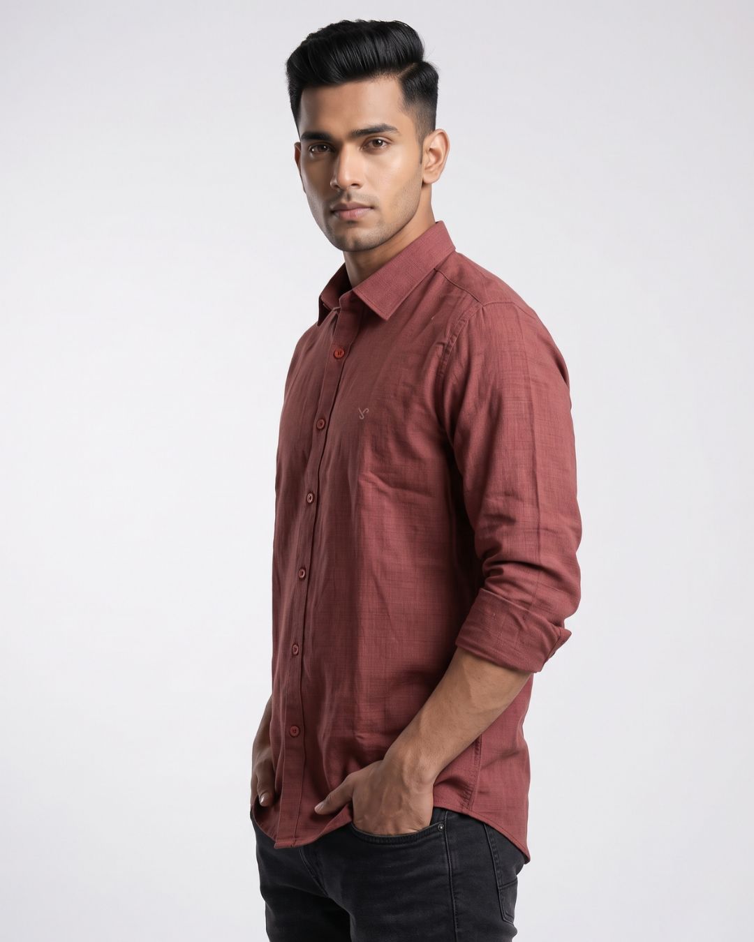 A man wearing a long-sleeve button-up shirt in a dusty rose or muted terracotta color, paired with dark jeans, standing against a plain light gray studio background.