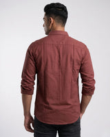 A man viewed from the back wearing a rust-colored, long-sleeve linen button-up shirt and dark jeans against a light gray studio background.