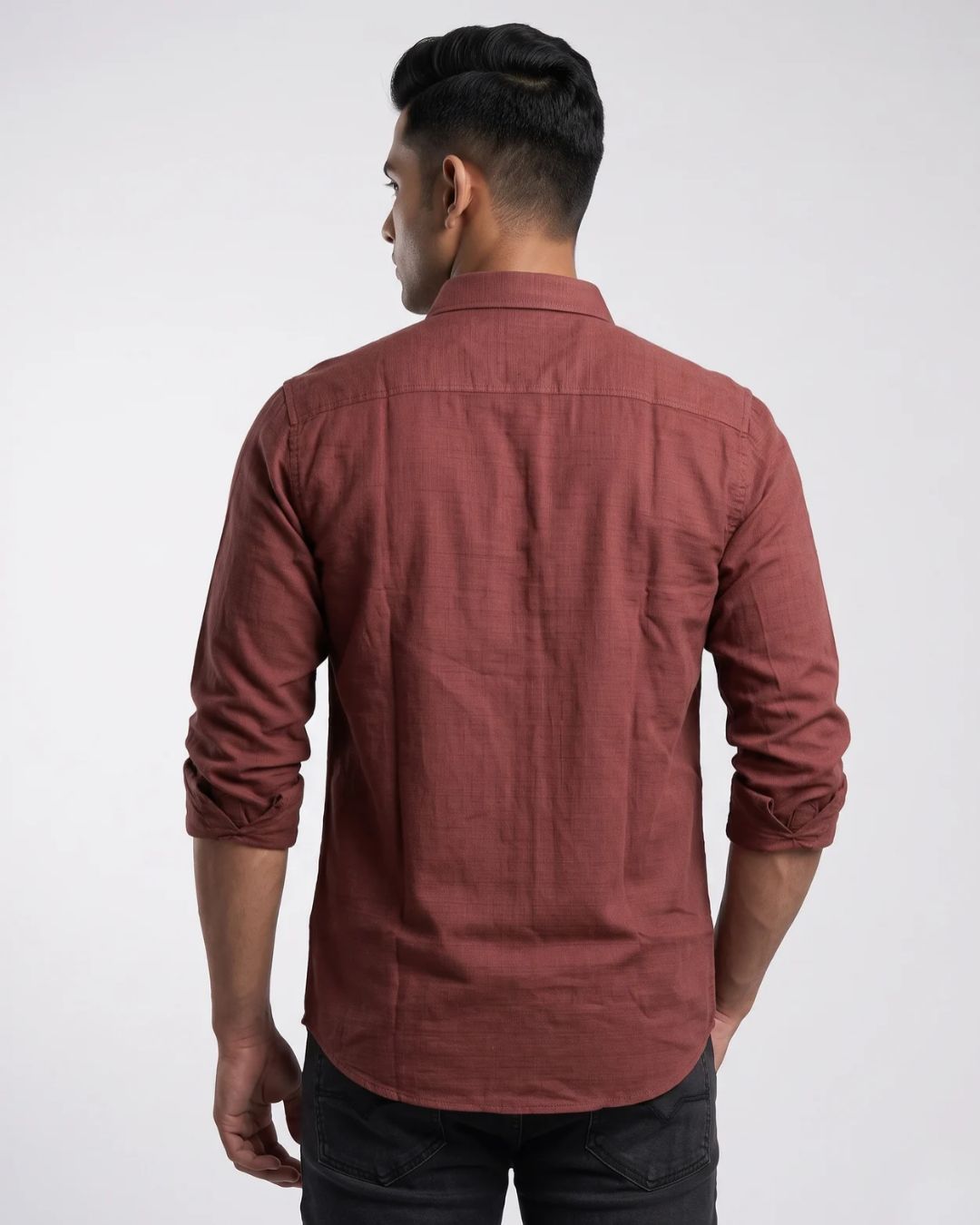 A man viewed from the back wearing a rust-colored, long-sleeve linen button-up shirt and dark jeans against a light gray studio background.