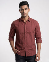 A man wearing a long-sleeve button-up shirt in a dusty rose or muted terracotta color, paired with dark jeans, standing against a light gray studio background.