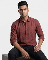 A man wearing a long-sleeve button-up shirt in a dusty rose or muted terracotta color, paired with black jeans, sitting against a plain light gray studio background.