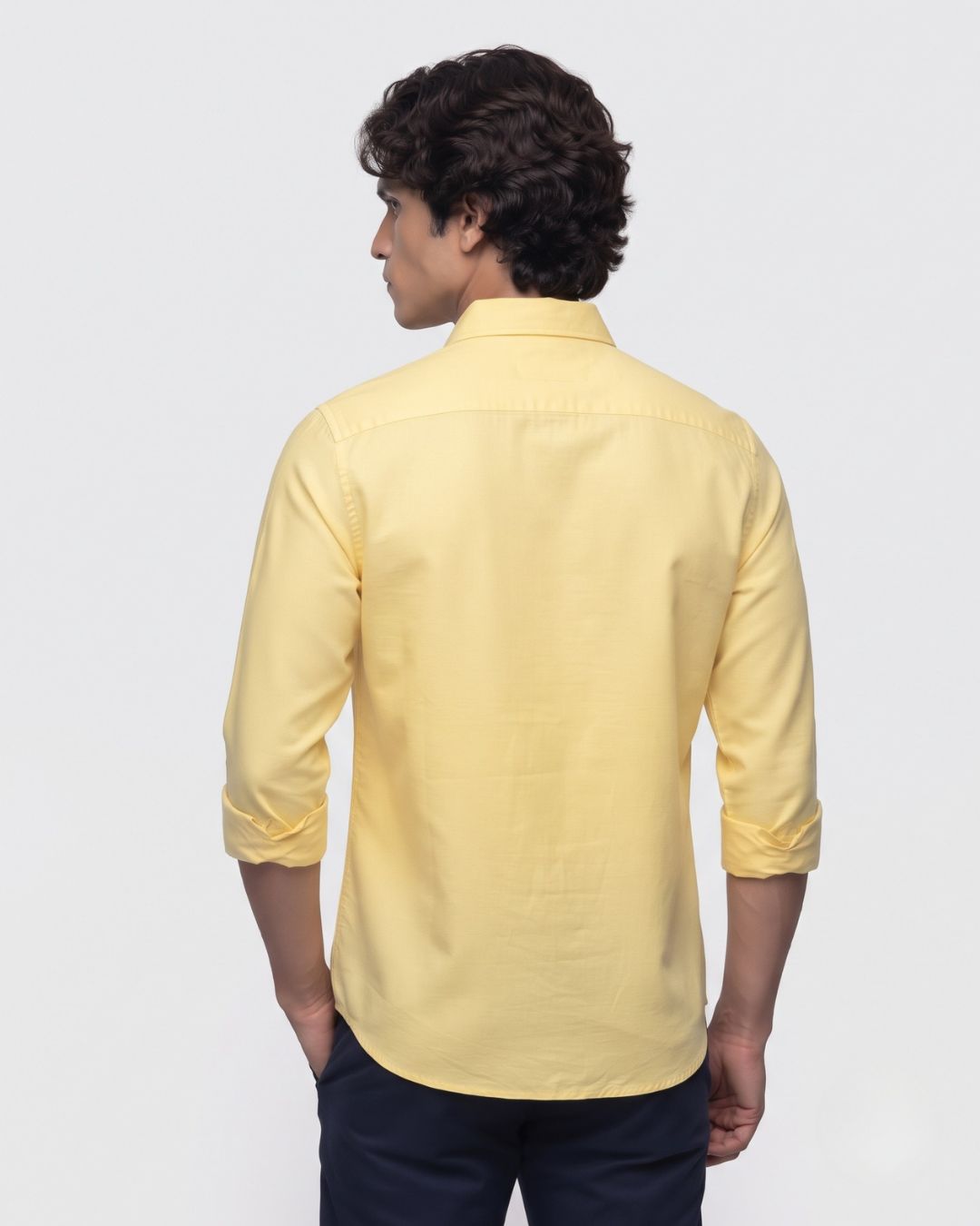 A man wearing a light yellow long-sleeve button-down shirt, shown from the back against a plain light gray studio background.