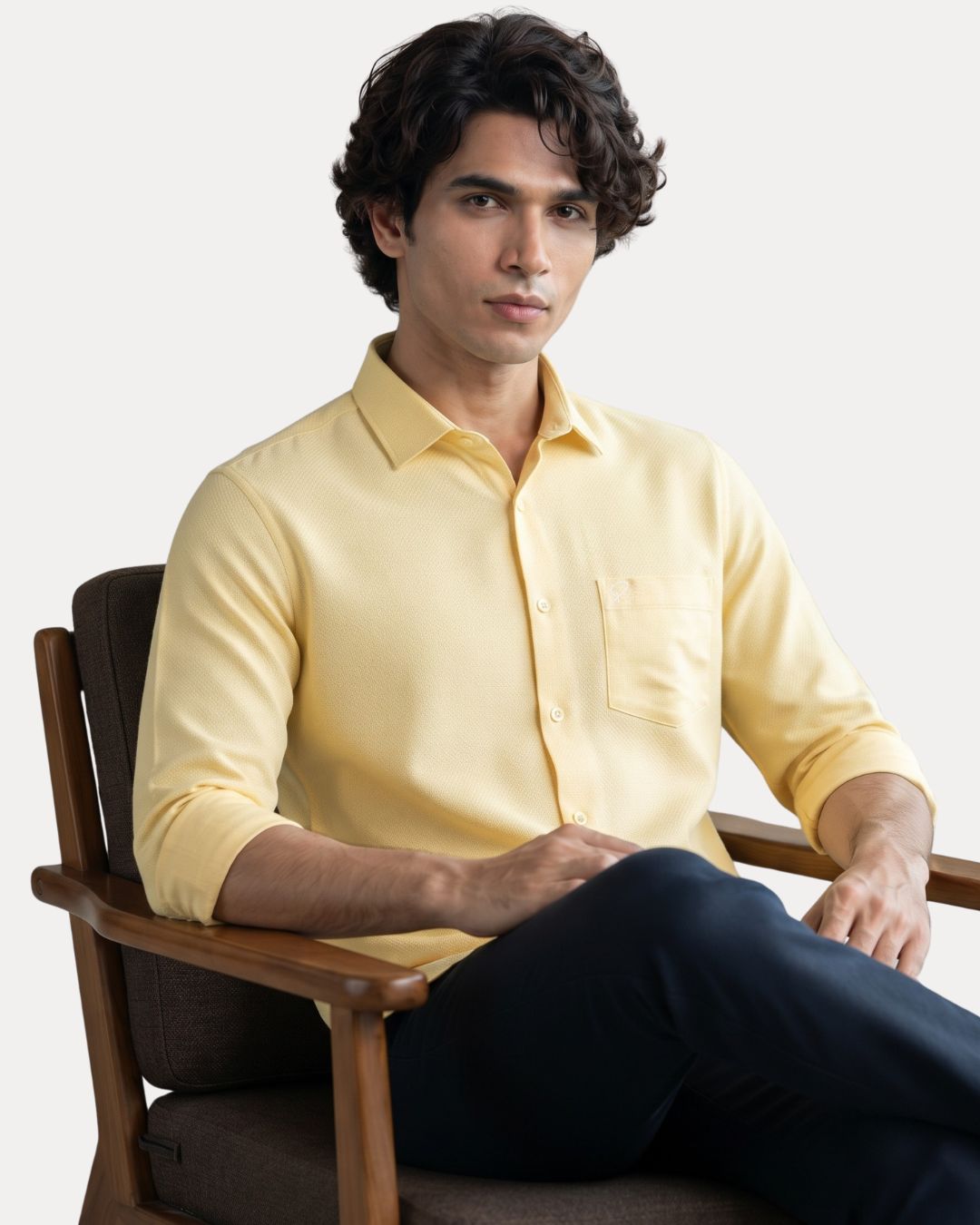 A man wearing a pale yellow long-sleeve button-up shirt and dark navy pants, seated in a wooden armchair against a light gray studio background.