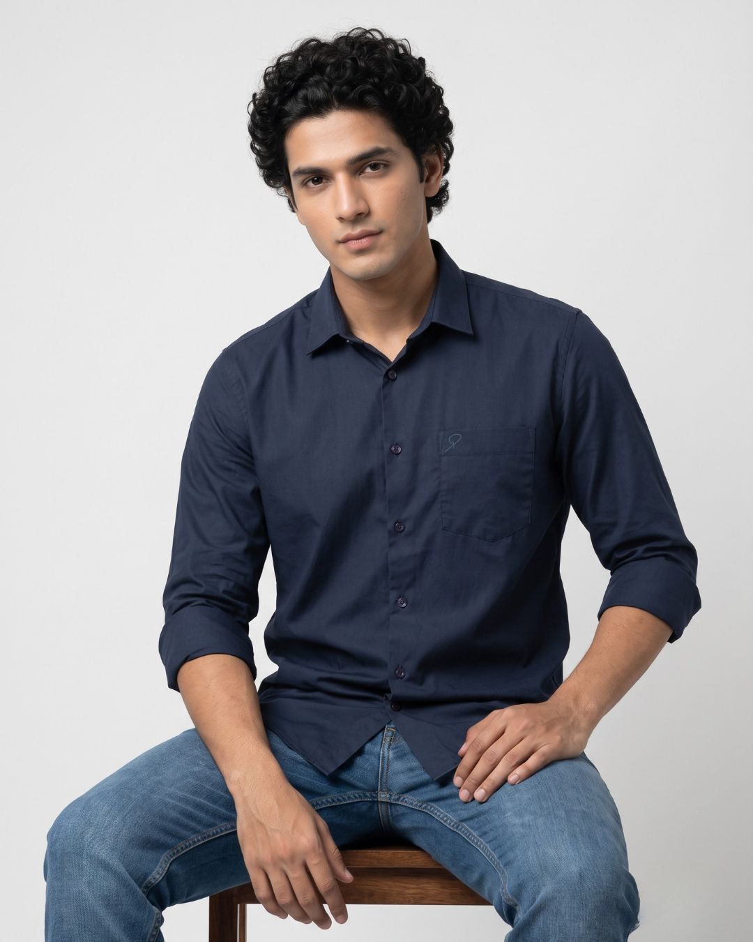 A man wearing a dark navy blue long-sleeve button-up shirt and blue jeans, sitting on a wooden stool against a light gray background.