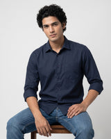 A man wearing a dark navy blue long-sleeve button-up shirt and blue jeans, sitting on a wooden stool against a light gray background.