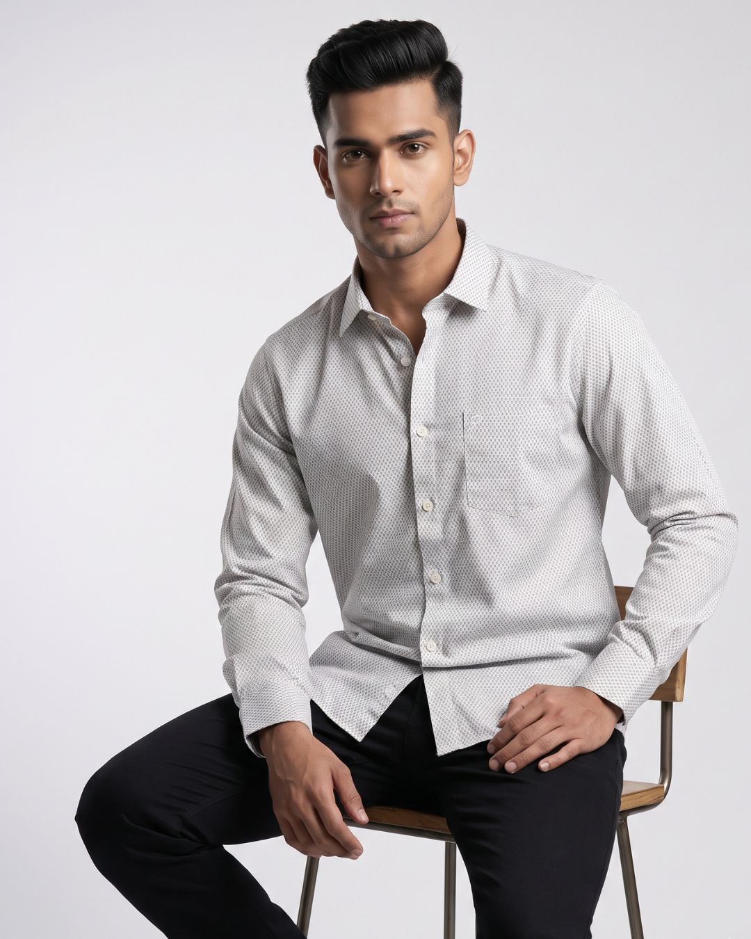 A man wearing a light gray long-sleeve button-down shirt with a subtle micro-pattern, paired with black trousers, sitting on a wooden chair against a light gray studio background.