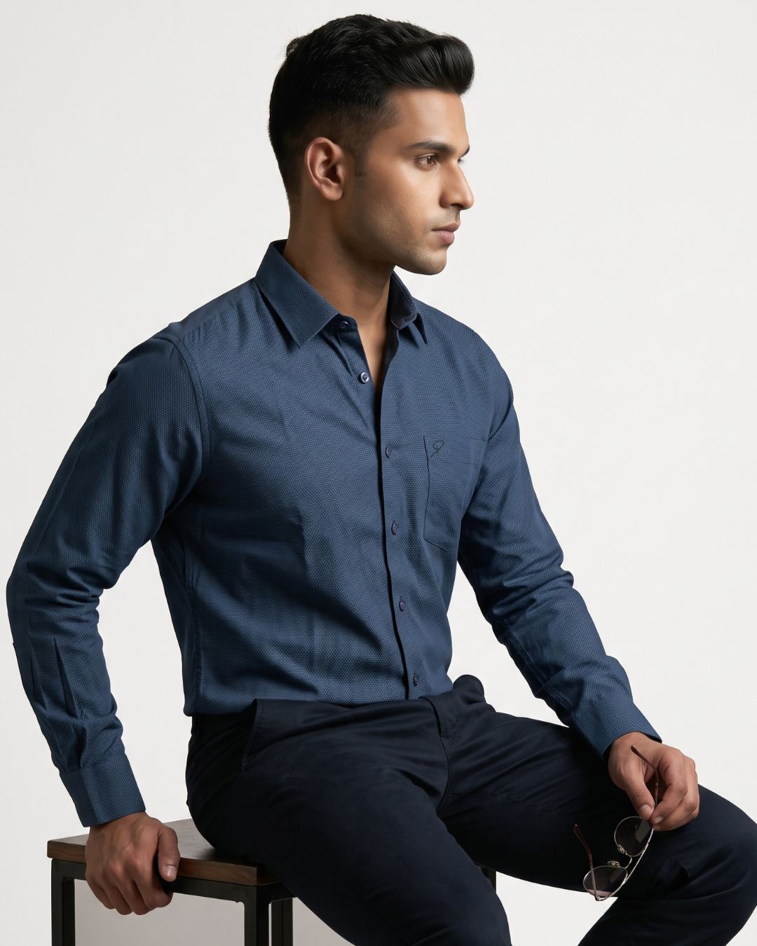 A man wearing a dark blue long-sleeve button-down shirt and dark trousers, sitting on a stool against a light gray background.