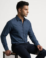 A man wearing a dark blue long-sleeve button-down shirt and dark trousers, sitting on a stool against a light gray background.
