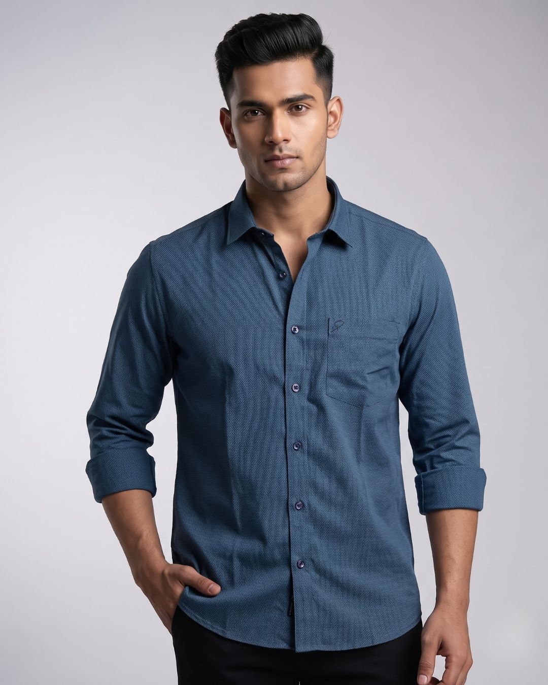 A man wearing a long-sleeve, button-down blue shirt with a subtle vertical stripe pattern, paired with black trousers, photographed against a neutral gray studio background.