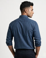 A man shown from the back wearing a navy blue long-sleeve button-down shirt and dark trousers, photographed against a clean light gray studio background.
