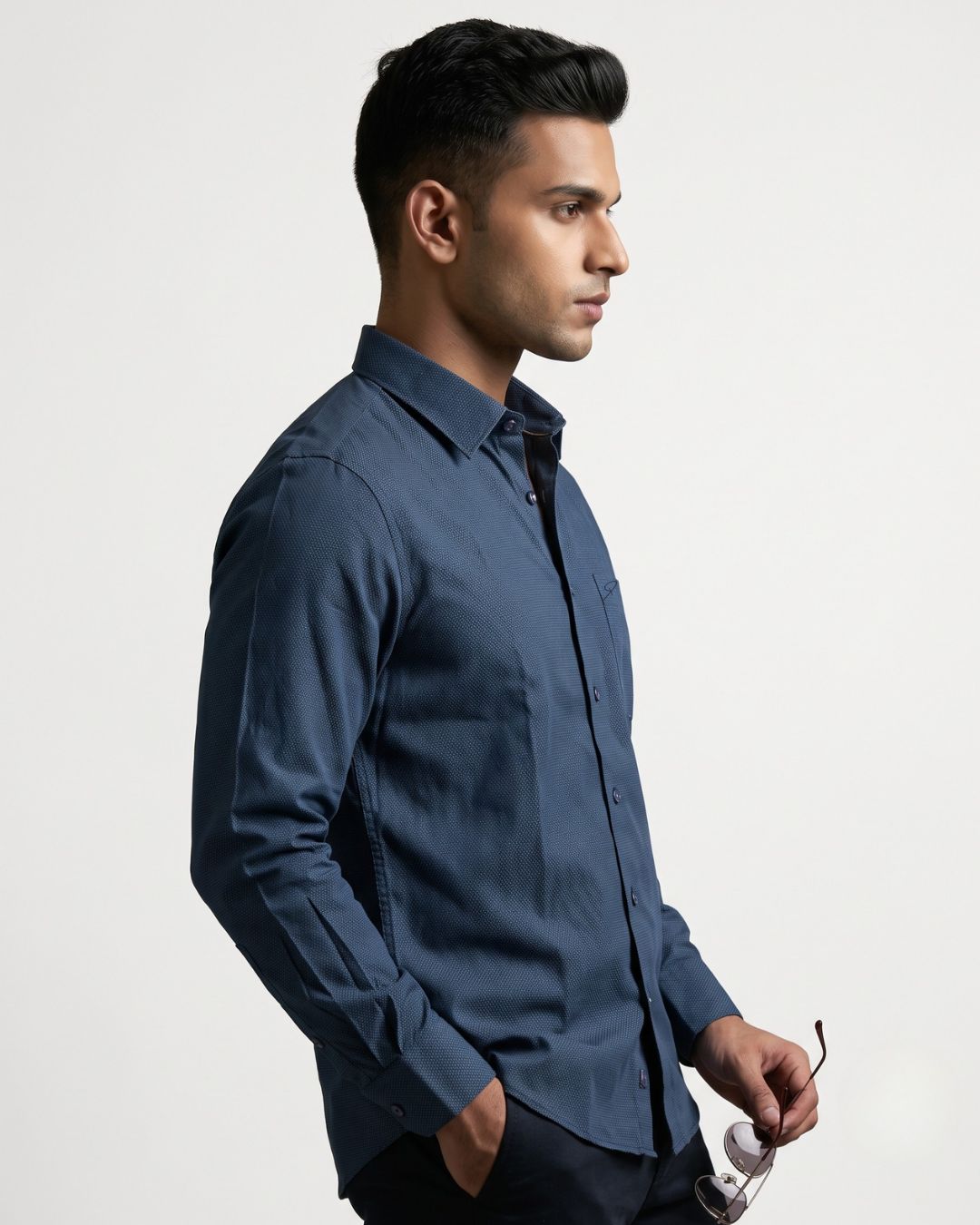 A man in profile wearing a dark blue long-sleeve button-down shirt, holding a pair of sunglasses against a plain light gray studio background.