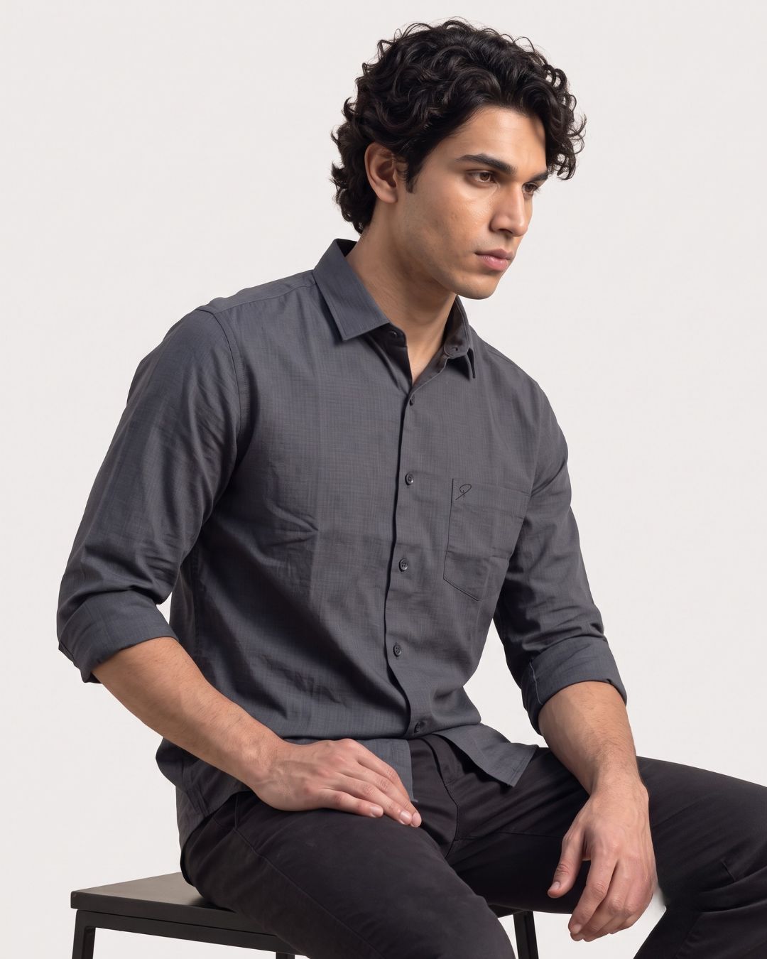 A man wearing a dark gray long-sleeve button-down shirt and black pants, sitting on a stool against a light gray studio background.
