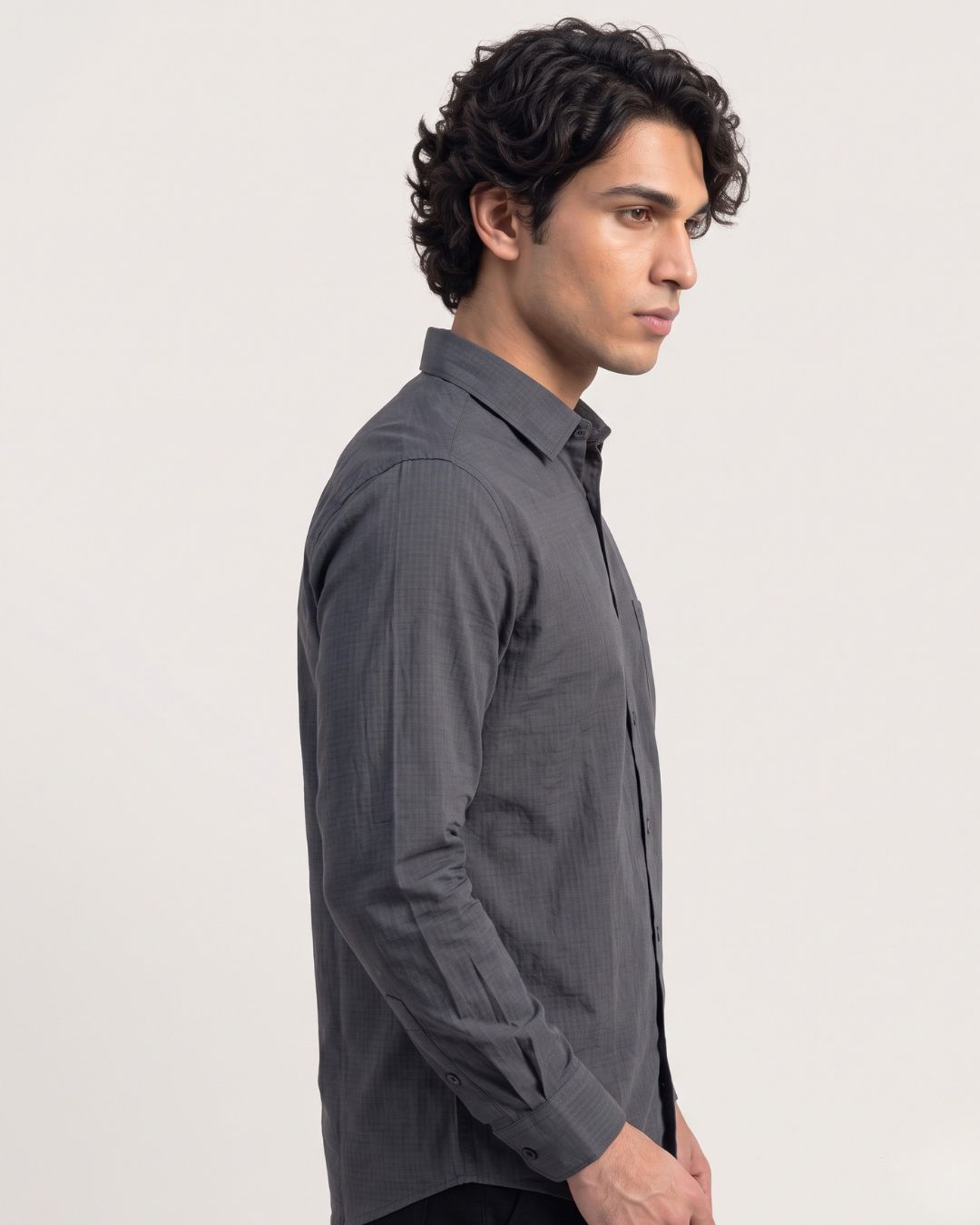 A man in side profile wearing a dark gray long-sleeve button-down shirt with a subtle grid pattern, photographed against a light gray studio background.