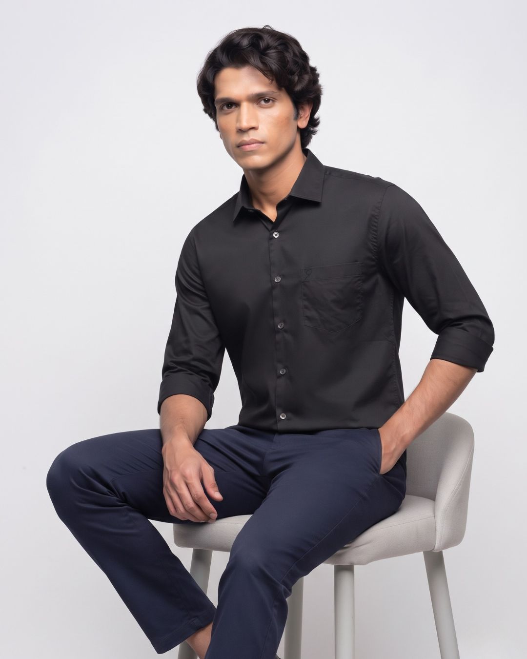 A man wearing a black long-sleeve button-up shirt and navy blue trousers, sitting on a light gray upholstered stool against a plain light gray studio background.