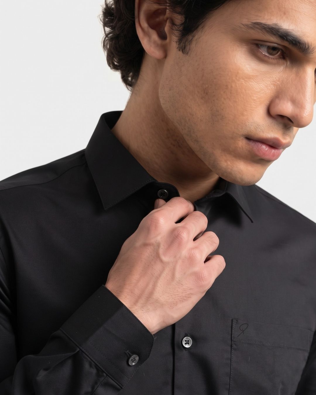 A close-up of a man wearing a black long-sleeve button-down dress shirt, adjusting the collar with his hand against a light gray background.