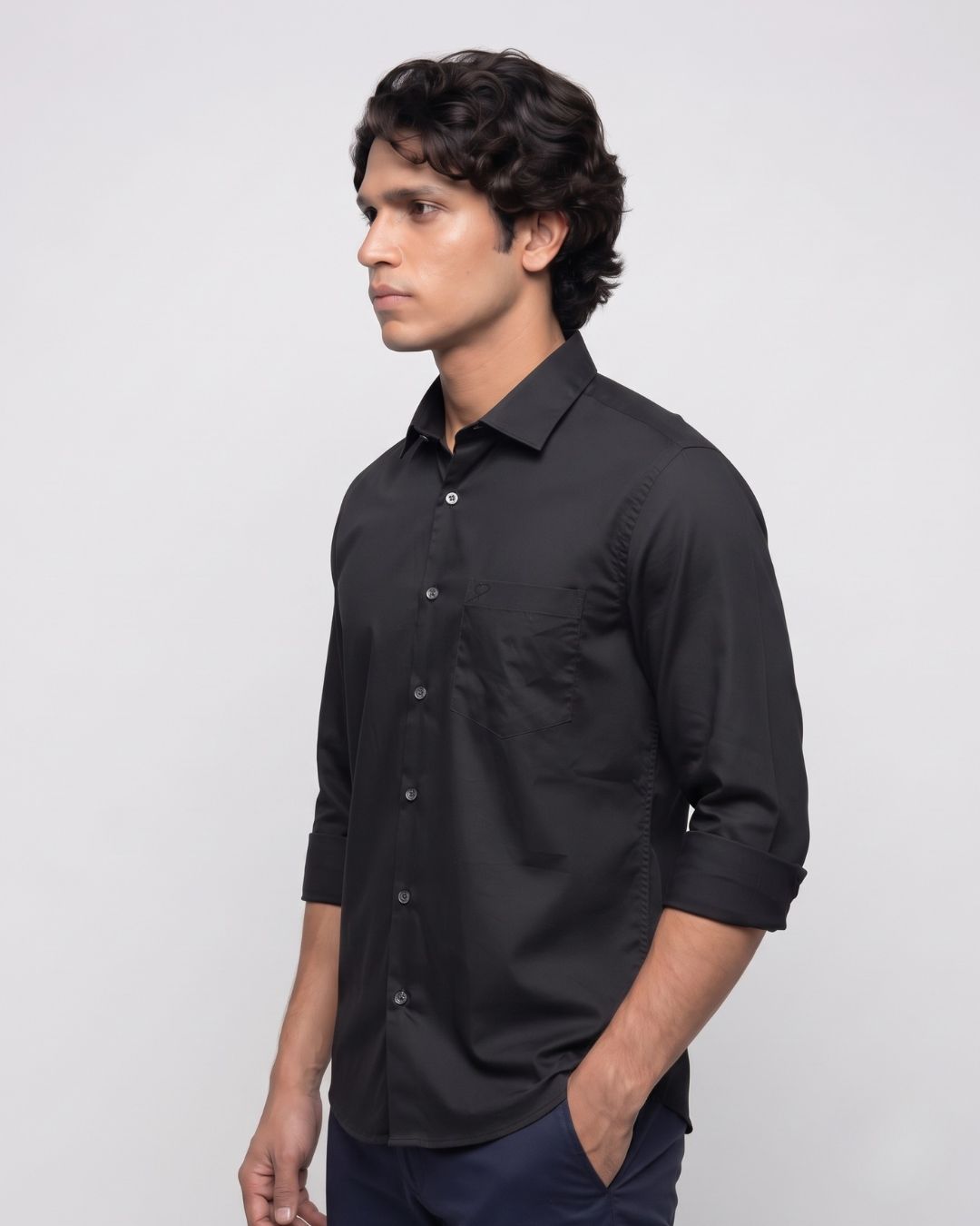 A man wearing a black long-sleeve button-down shirt with rolled-up sleeves, paired with dark trousers, standing against a light gray studio background.