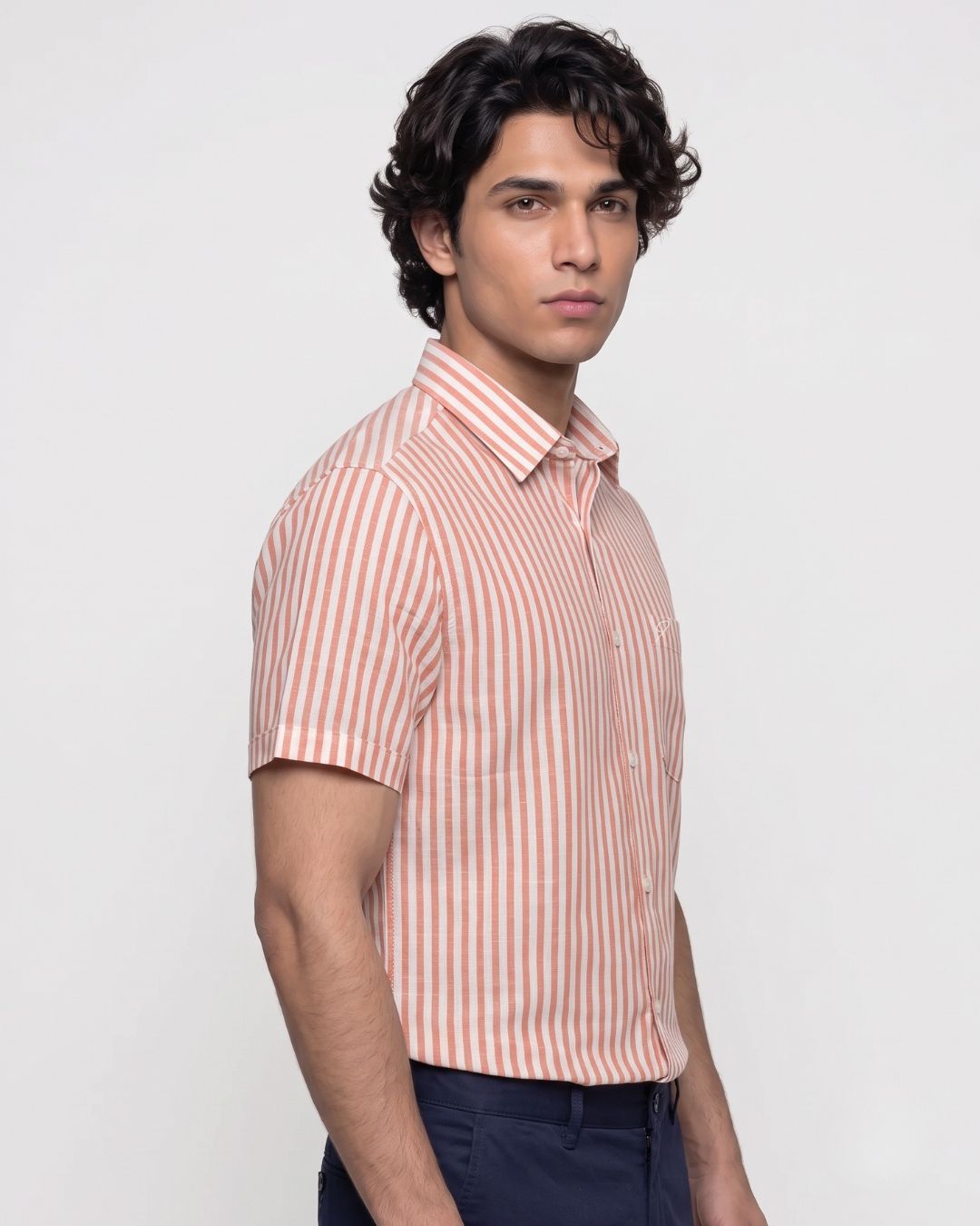 A man wearing a short-sleeve button-up shirt with vertical peach and white stripes, paired with dark navy trousers, photographed against a light gray studio background.