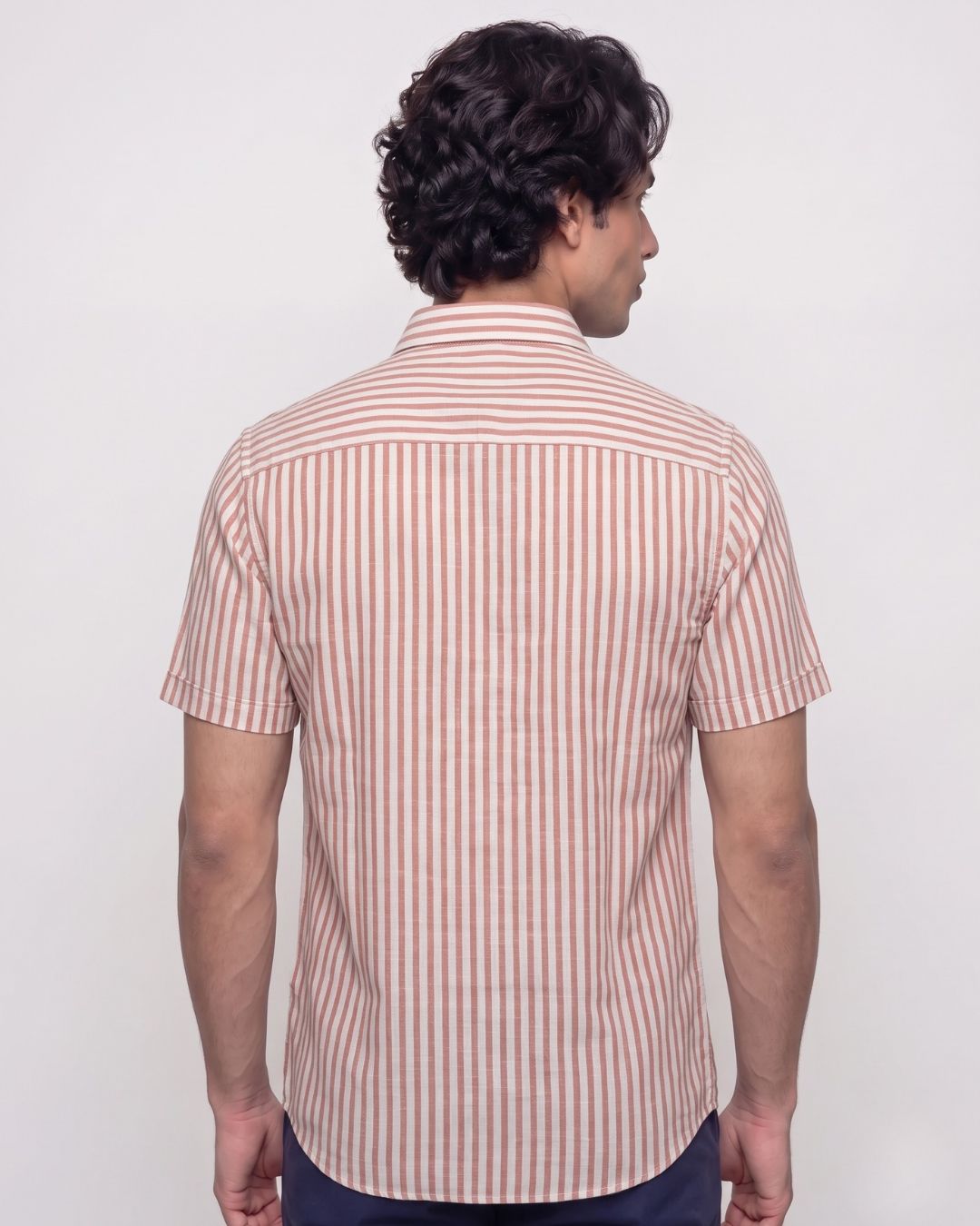 A man viewed from the back wearing a short-sleeve button-up shirt with vertical pink and white stripes, paired with dark navy pants, against a plain light gray background.