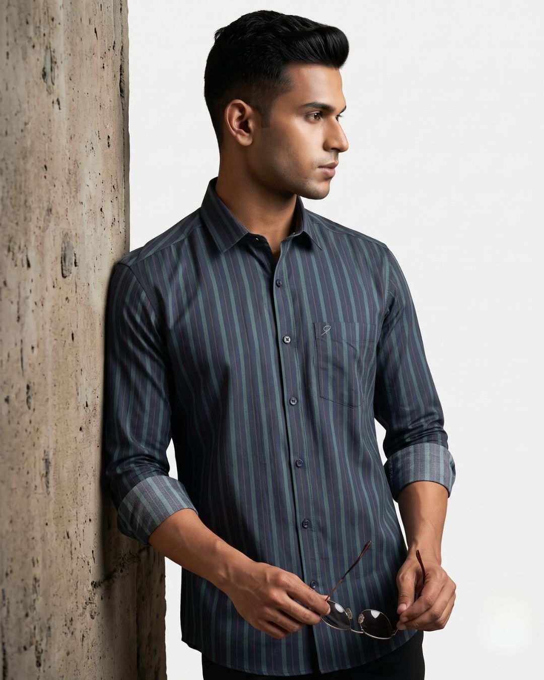 A man wearing a dark gray and black vertically striped button-down shirt leans against a textured concrete pillar, holding a pair of sunglasses.