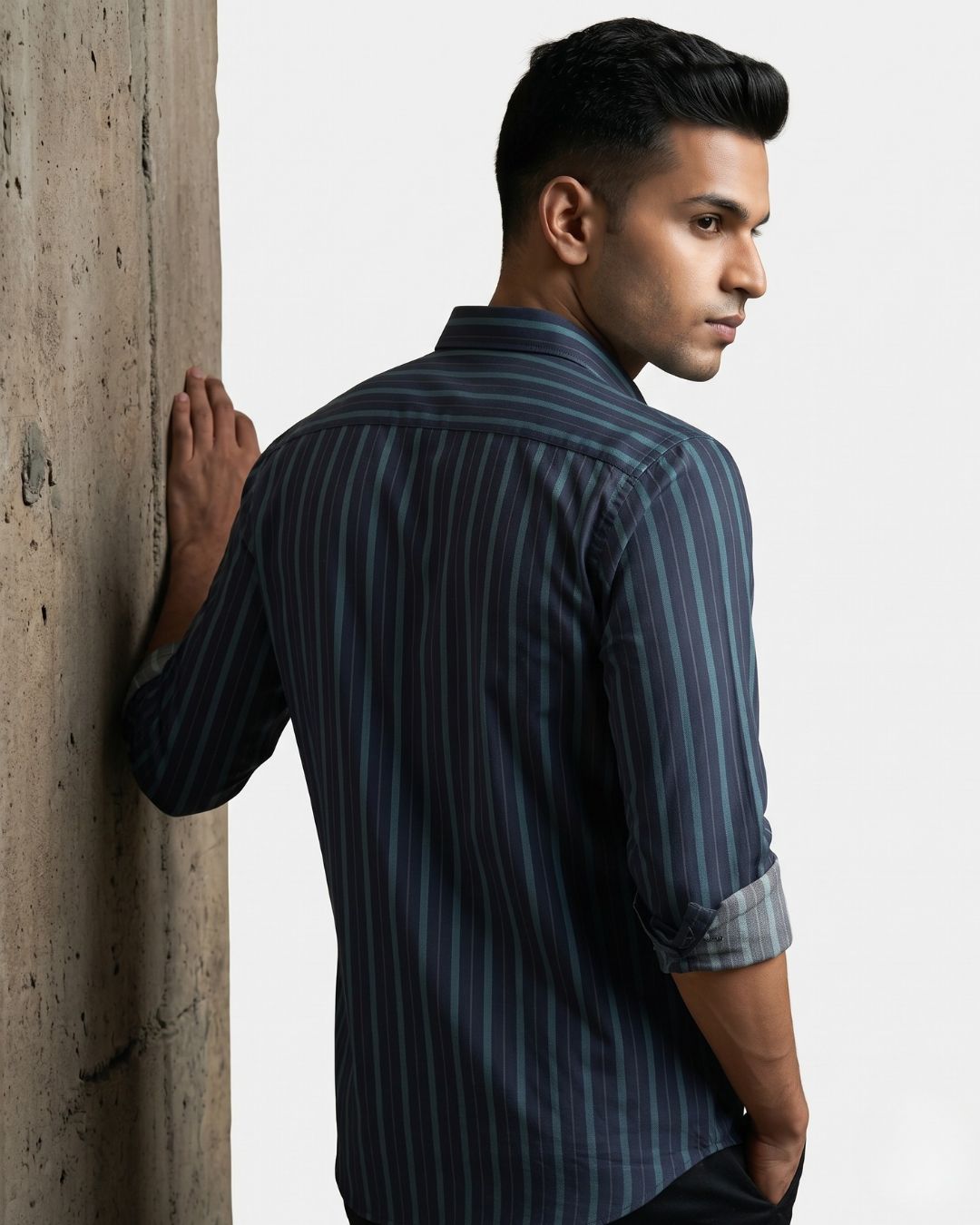 A man wearing a dark navy blue and black vertically striped button-down shirt, leaning against a textured concrete pillar against a white studio background.