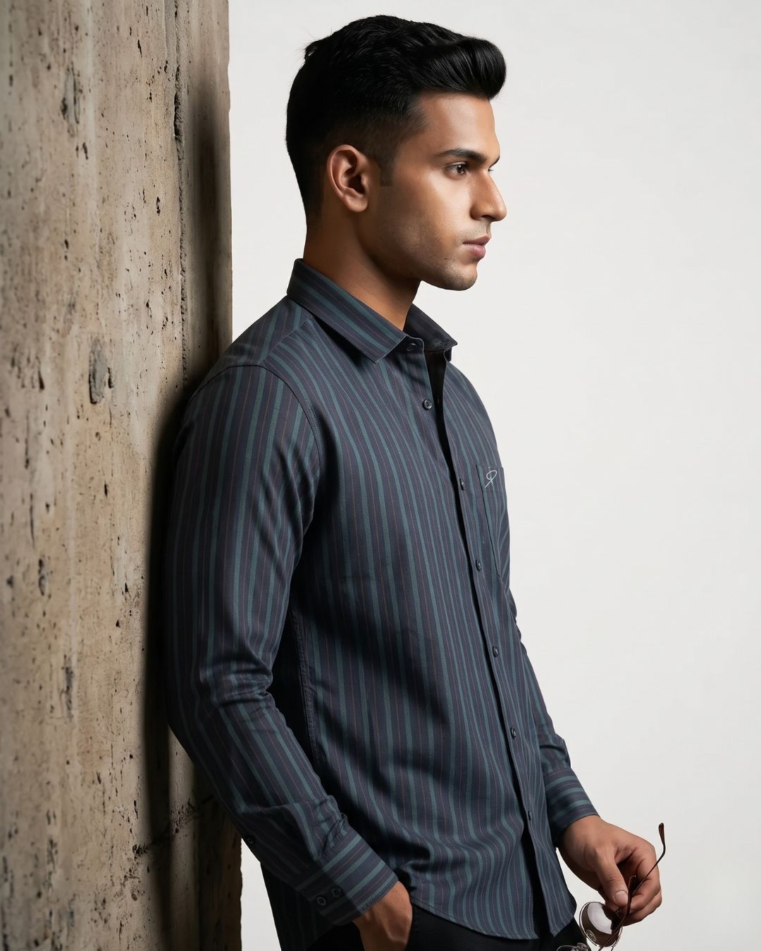 A man wearing a dark gray and black vertically striped long-sleeve button-down shirt, leaning against a textured concrete pillar.