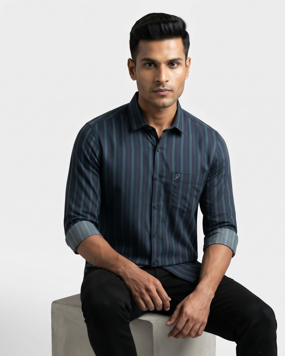 A man wearing a dark navy blue and black vertically striped long-sleeve button-down shirt, sitting on a light gray block against a plain light gray background.