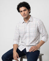 A man wearing a white long-sleeve button-up shirt with a subtle blue and pink grid pattern, paired with dark blue jeans, sitting on a stool against a light gray background.