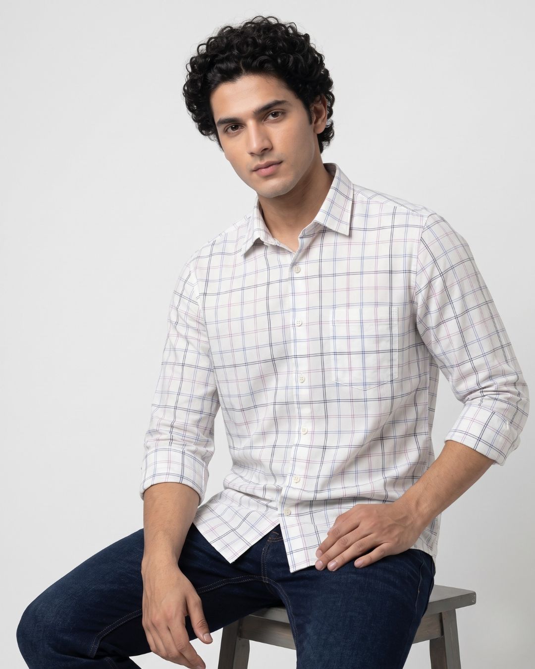 A man wearing a white long-sleeve button-up shirt with a subtle blue and pink grid pattern, paired with dark blue jeans, sitting on a stool against a light gray background.