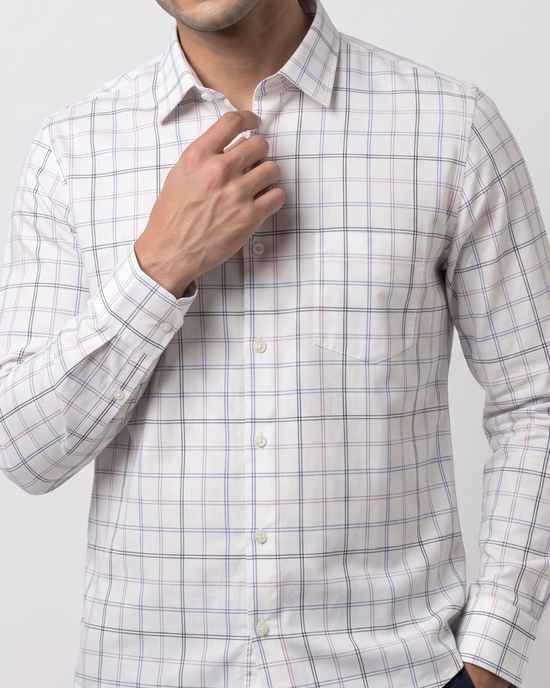 A man wearing a long-sleeve button-down shirt with a white base and a subtle pink and blue windowpane plaid pattern. He is adjusting the collar with one hand, shown from the chest down against a light gray studio background.
