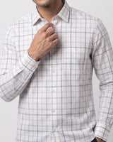 A man wearing a long-sleeve button-down shirt with a white base and a subtle pink and blue windowpane plaid pattern. He is adjusting the collar with one hand, shown from the chest down against a light gray studio background.