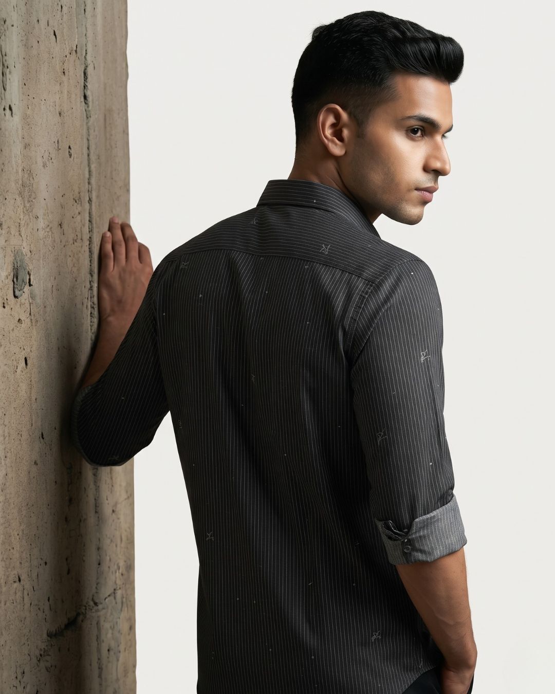 A man wearing a dark gray pinstriped shirt with subtle paint splatter details, leaning against a textured concrete pillar against a light gray background.