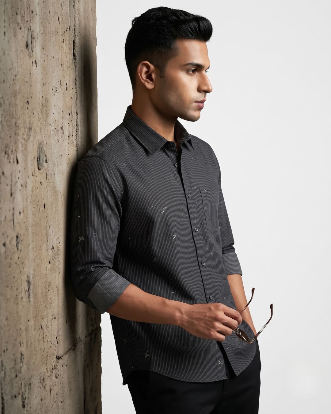A man wearing a dark gray long-sleeve button-down shirt with a subtle micro-pattern, leaning against a textured concrete pillar next to a plain white wall.