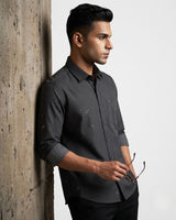 A man wearing a dark gray long-sleeve button-down shirt with a subtle micro-pattern, leaning against a textured concrete pillar next to a plain white wall.