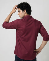 A man wearing a burgundy long-sleeve button-down shirt, shown from the back and side against a plain light gray studio background.