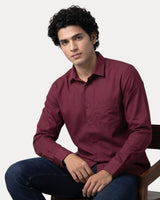 A man wearing a long-sleeve burgundy button-down shirt and dark blue jeans, sitting on a wooden chair against a light gray background.