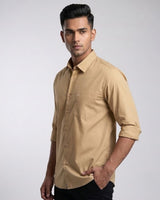 A man wearing a tan long-sleeve button-up shirt with rolled-up sleeves and black pants, standing against a light gray studio background.