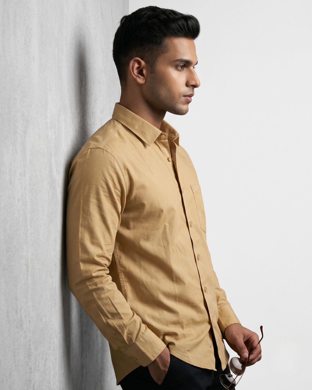 A man wearing a tan long-sleeve button-down shirt leans against a light gray textured wall, holding a pair of sunglasses.