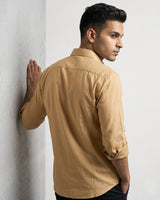A man wearing a tan long-sleeve linen shirt, leaning against a textured gray wall in a studio setting.