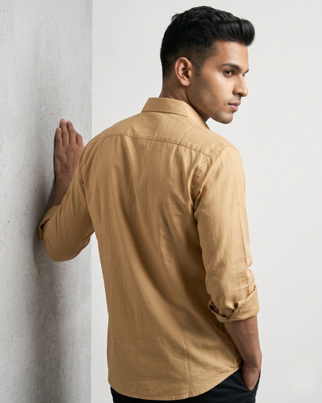 A man wearing a tan long-sleeve linen shirt, leaning against a textured gray wall in a studio setting.