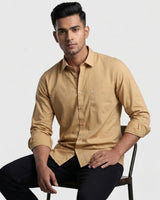 A man wearing a tan long-sleeve button-up shirt and black pants, seated on a dark chair against a light gray studio background.