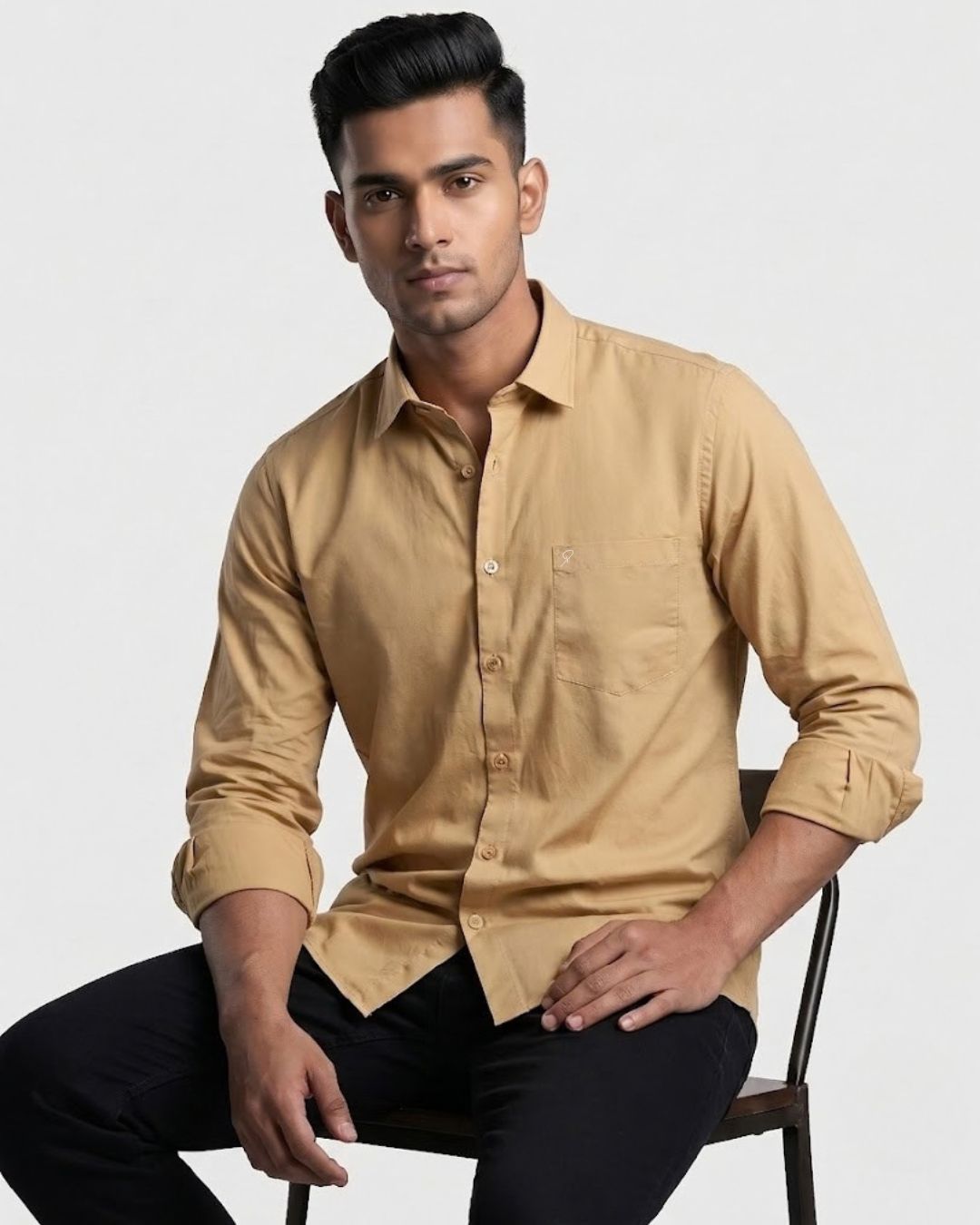 A man wearing a tan long-sleeve button-up shirt and black pants, seated on a dark chair against a light gray studio background.
