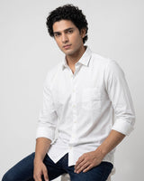 A man wearing a white long-sleeve button-up shirt with rolled-up sleeves and dark blue jeans, seated against a plain light gray studio background.